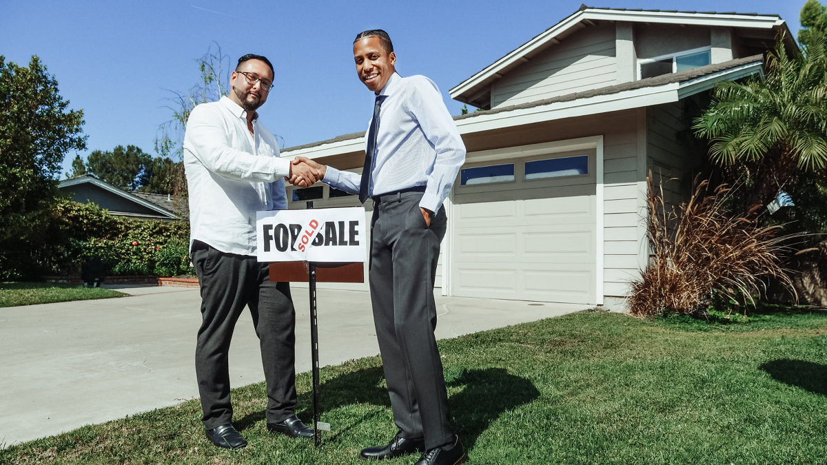 Real estate broker and new house owner shaking hands in front of sold signboard