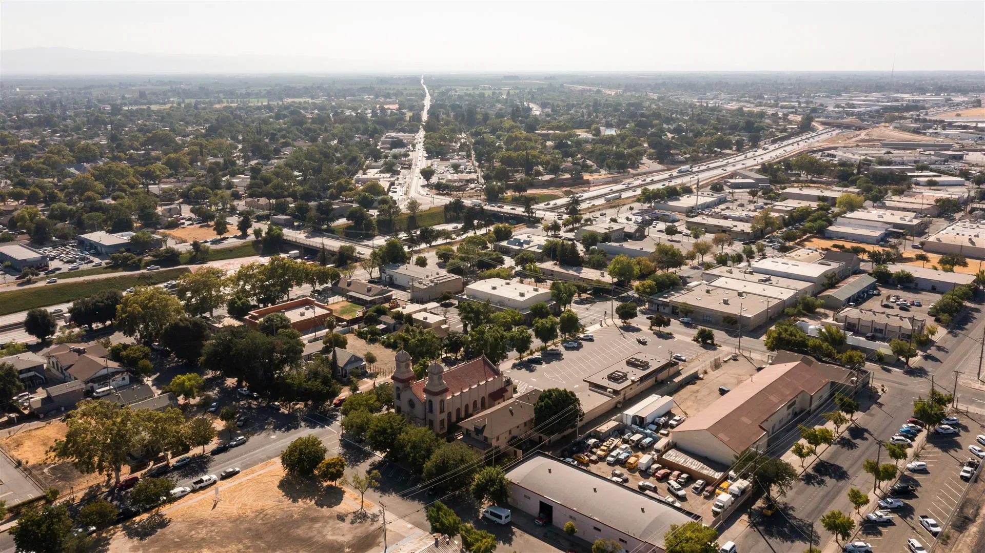 Aerial view of a sprawling city with dense greenery, roads, and varied buildings under a hazy sky. Prominent church with two towers in the foreground.