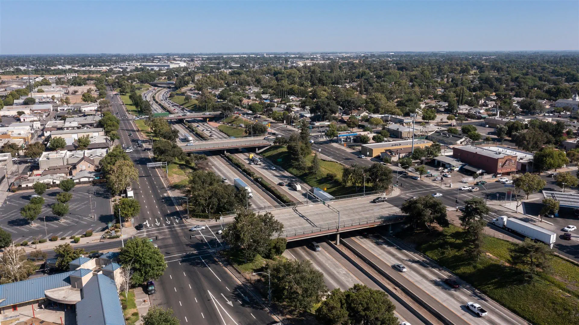 Aerial view of a busy urban highway interchange surrounded by trees and buildings under a clear blue sky, conveying a sense of movement and urban life.