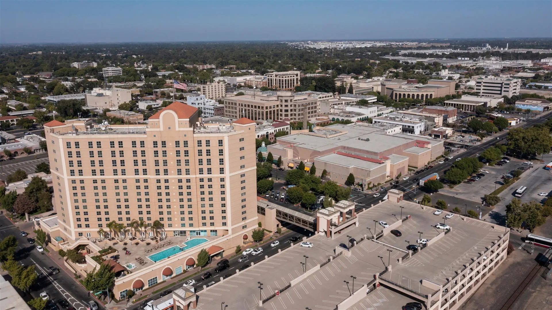 Aerial view of a tall beige hotel with a rooftop pool surrounded by trees. The urban landscape shows a mix of buildings and greenery under a clear sky.