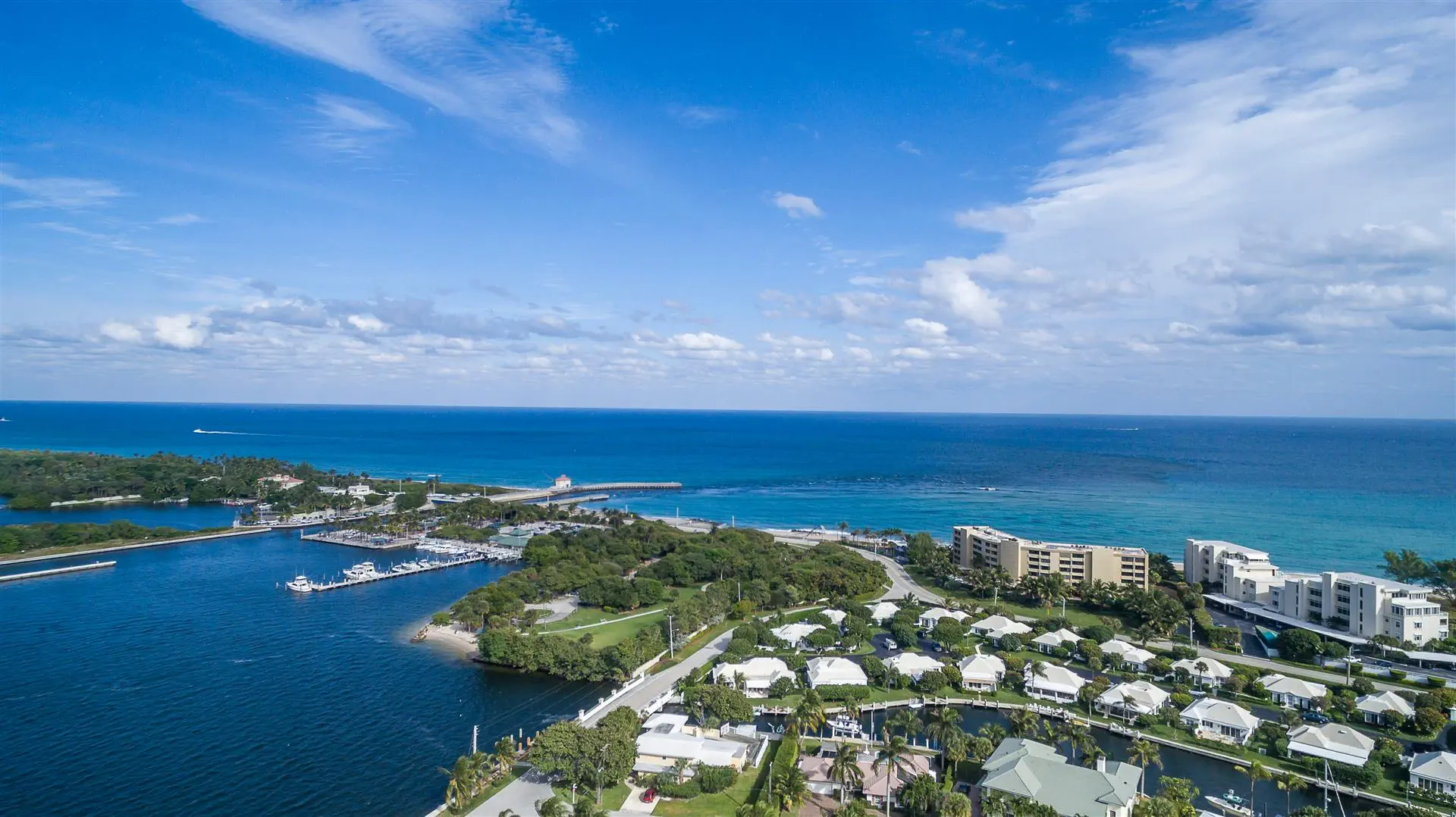 Aerial view of a coastal town with white-roofed houses, a marina, and lush greenery against a backdrop of blue ocean and a cloudy sky. Peaceful vibe.