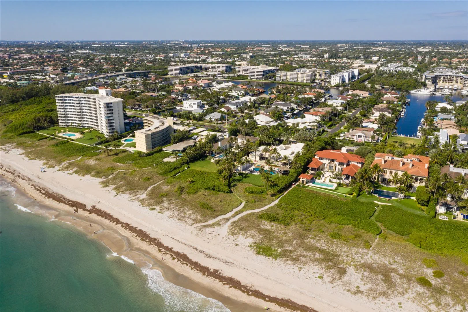 Aerial view of a coastal city with luxury beachfront homes, green lawns, and a sandy shoreline. High-rise buildings and a canal are visible in the background.