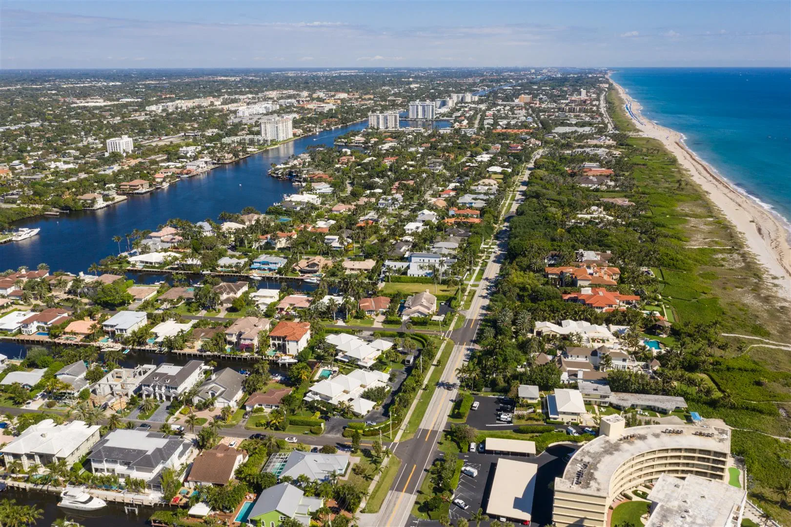 Aerial view of a coastal city with a river, lush greenery, and white buildings. The beach and ocean stretch along the right, conveying a serene, sunny atmosphere.