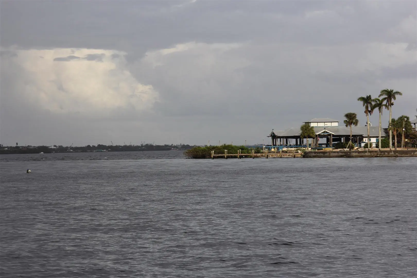 Cloudy sky over a calm waterfront with a small building surrounded by palm trees on the right. The scene conveys a serene, coastal atmosphere.