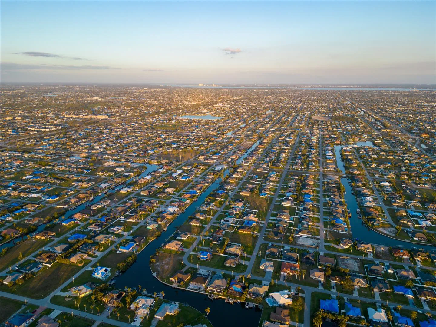 Aerial view of a sprawling suburban landscape with neat rows of houses and winding waterways. The blue sky and warm lighting create a serene atmosphere.