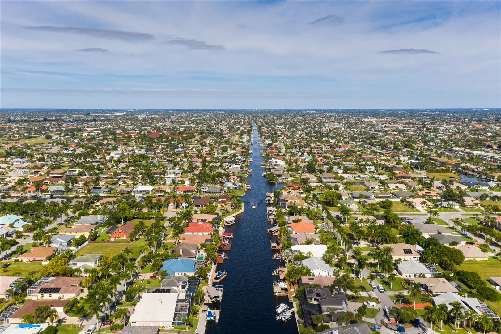 Aerial view of a coastal neighborhood with a central canal stretching to the horizon. Houses with various roof colors line the canal, amidst lush greenery.