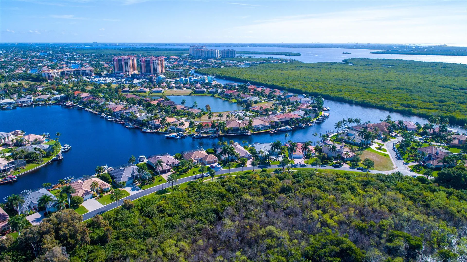 Aerial view of a coastal residential area with curved waterways, lush greenery, and clustered houses. High-rise buildings and a vast body of water in the background.