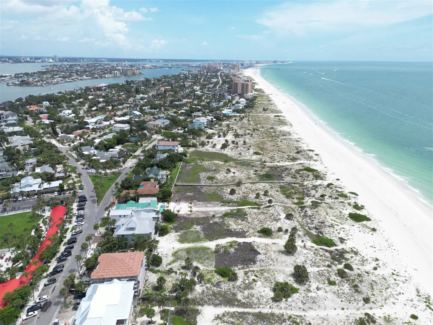 Aerial view of a coastal town with a wide beach and turquoise ocean on the right. Lush green vegetation and houses line the shore under a clear blue sky.