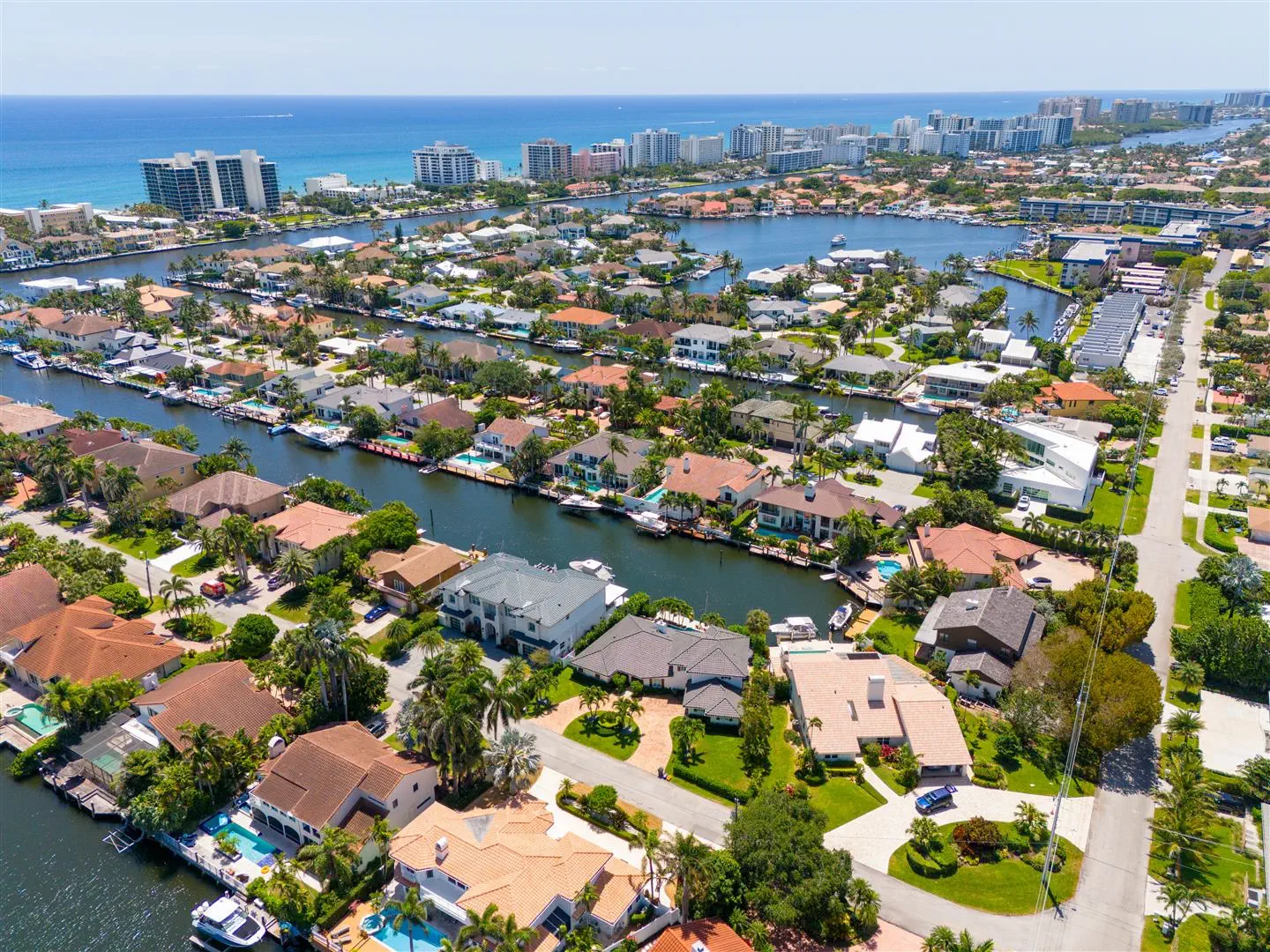 Aerial view of a coastal Florida neighborhood with canals between rows of houses. Lush greenery surrounds the homes, with a clear ocean in the background.