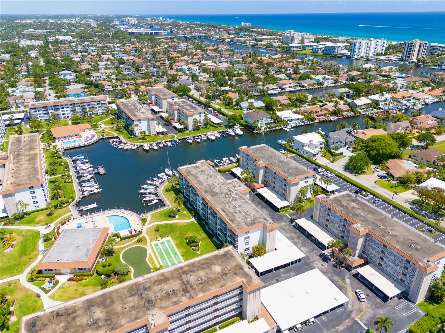 Aerial view of a coastal residential area with canals and boats. Tall apartments surround a large pool, athletic courts, and green spaces. Bright, sunny day.