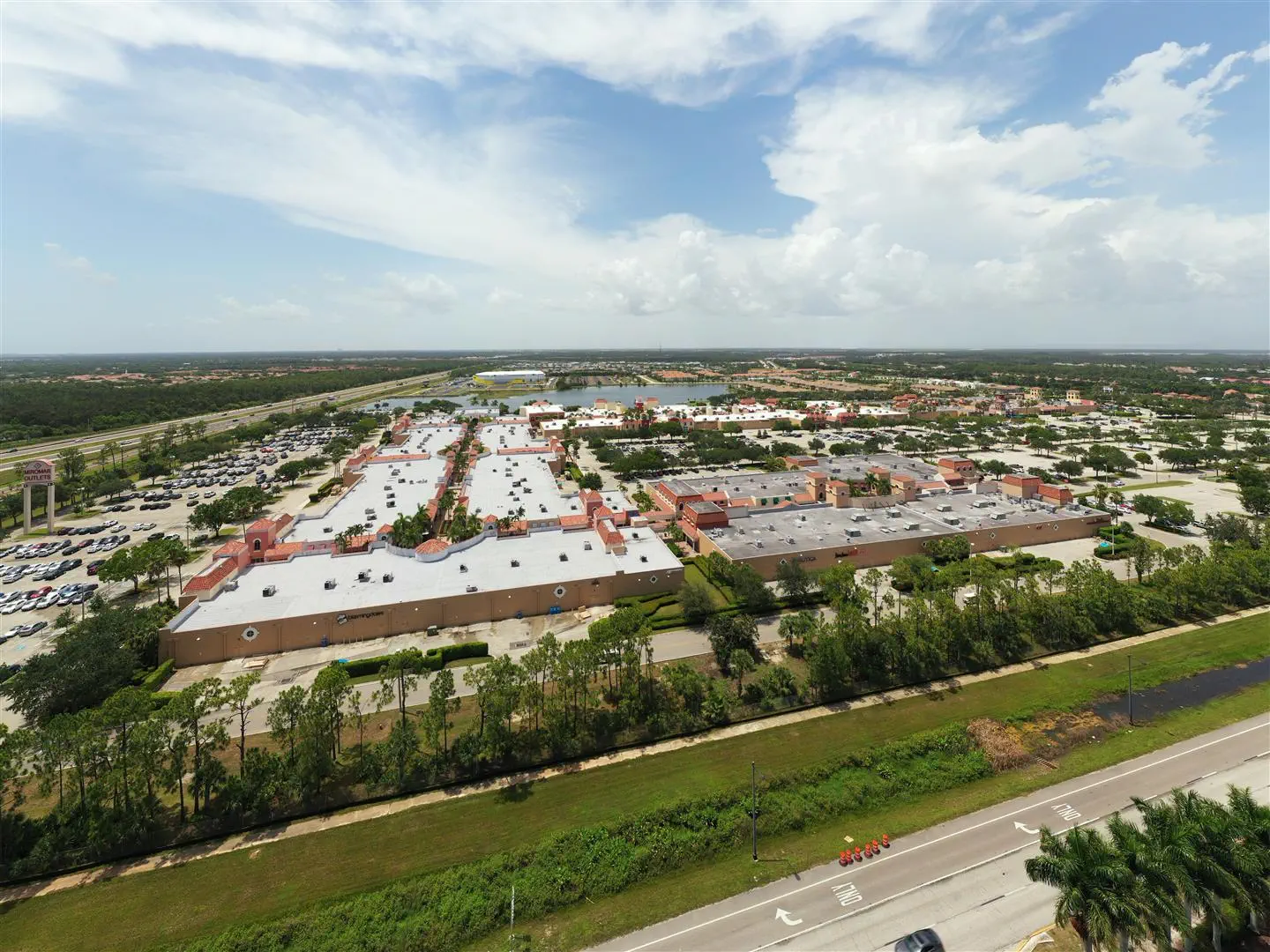 Aerial view of a large outlet shopping complex with red-tiled roofs, surrounded by parking lots, greenery, and roads under a partly cloudy sky.