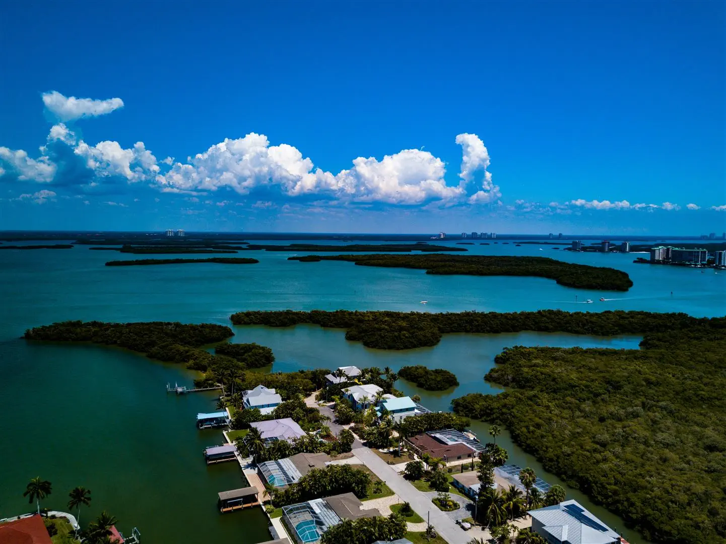 Aerial view of a coastal neighborhood with houses surrounded by lush green vegetation and waterways. The sky is clear with fluffy clouds, creating a serene and picturesque atmosphere.