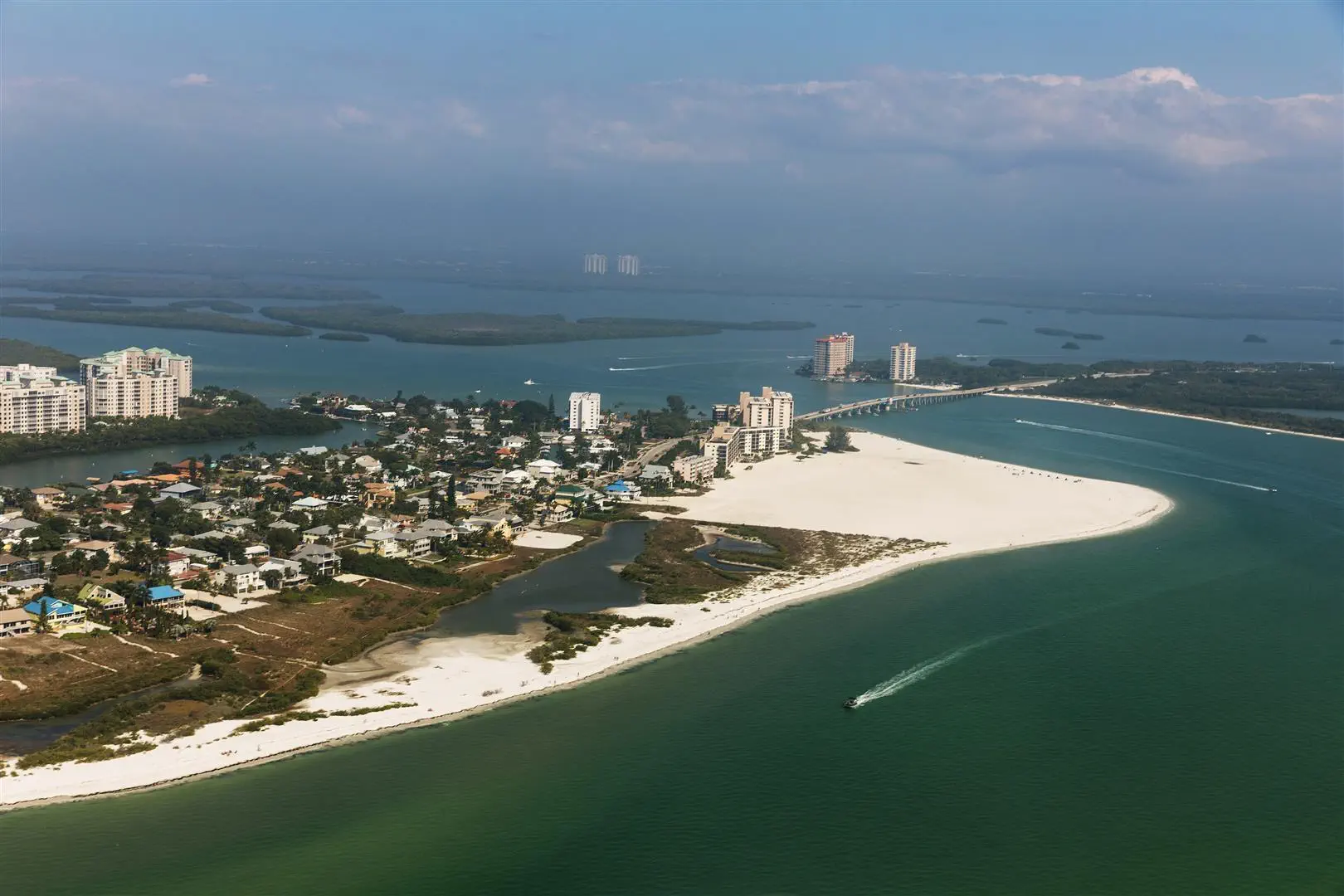 Aerial view of a coastal city with white sandy beaches and turquoise waters. High-rise buildings line the shoreline, while a boat leaves a wake in the sea.
