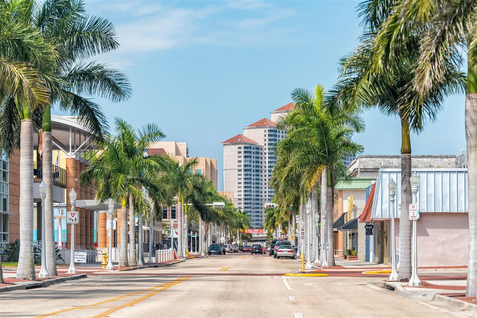 A sunny street flanked by tall palm trees and colorful low-rise buildings leads to distant high-rises. The scene conveys a tropical, vibrant atmosphere.