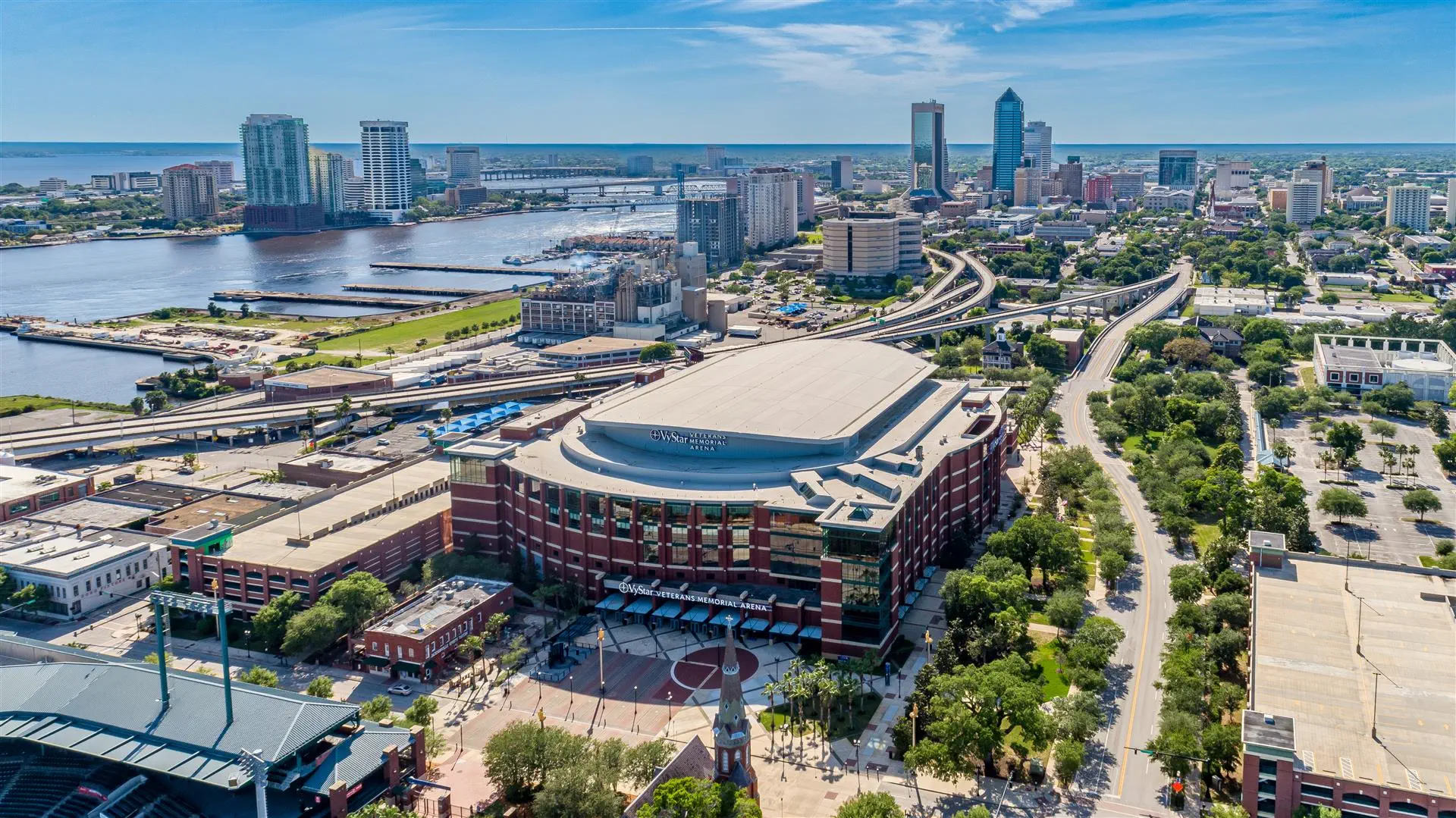 Aerial view of downtown Jacksonville, Florida, featuring high-rise buildings, a winding river, and a large stadium in the foreground under a clear blue sky.