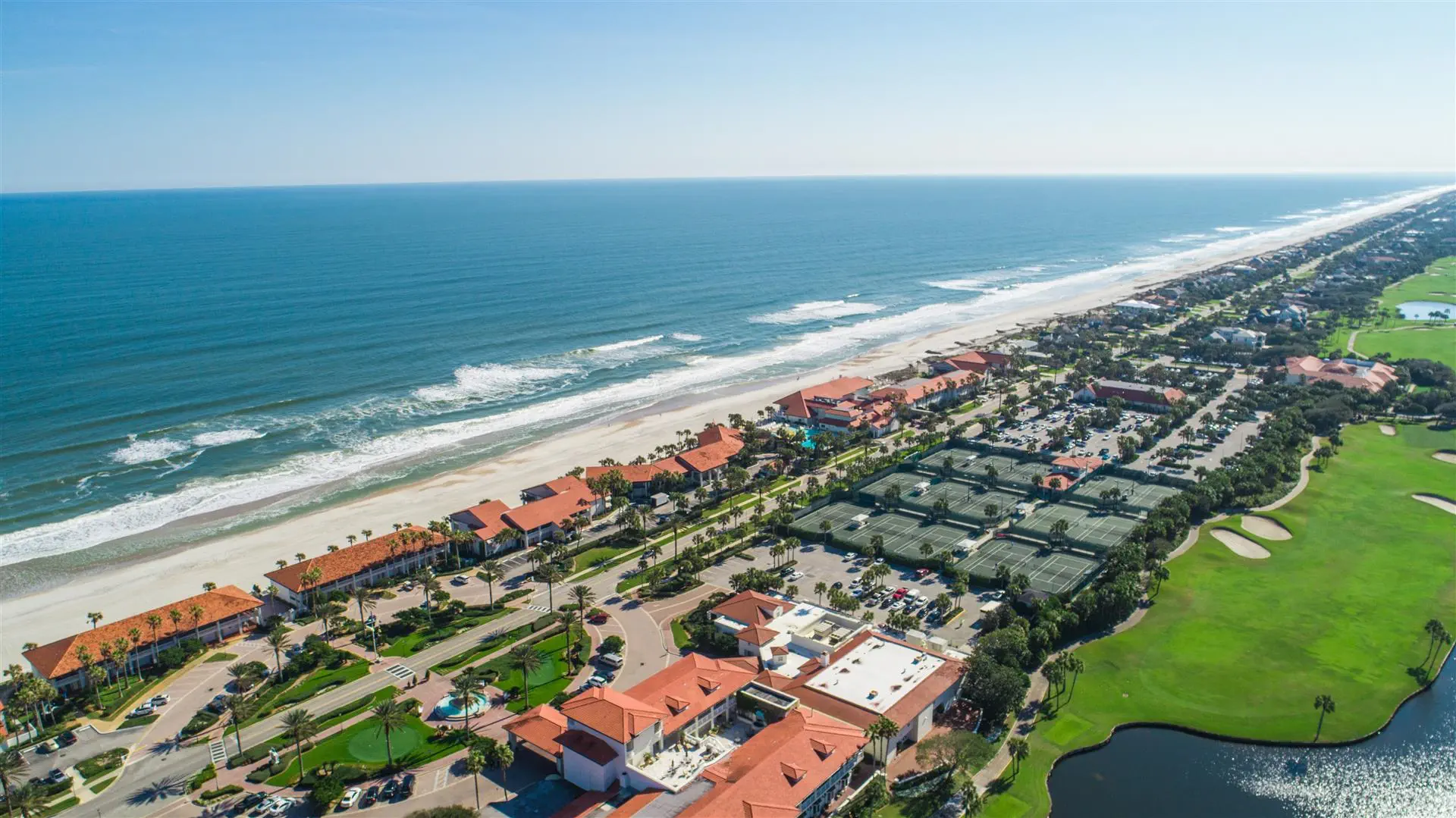 Aerial view of a coastal resort with red-roofed buildings lining a sandy beach. Tennis courts are visible, with the ocean and green golf course nearby. Bright and serene.