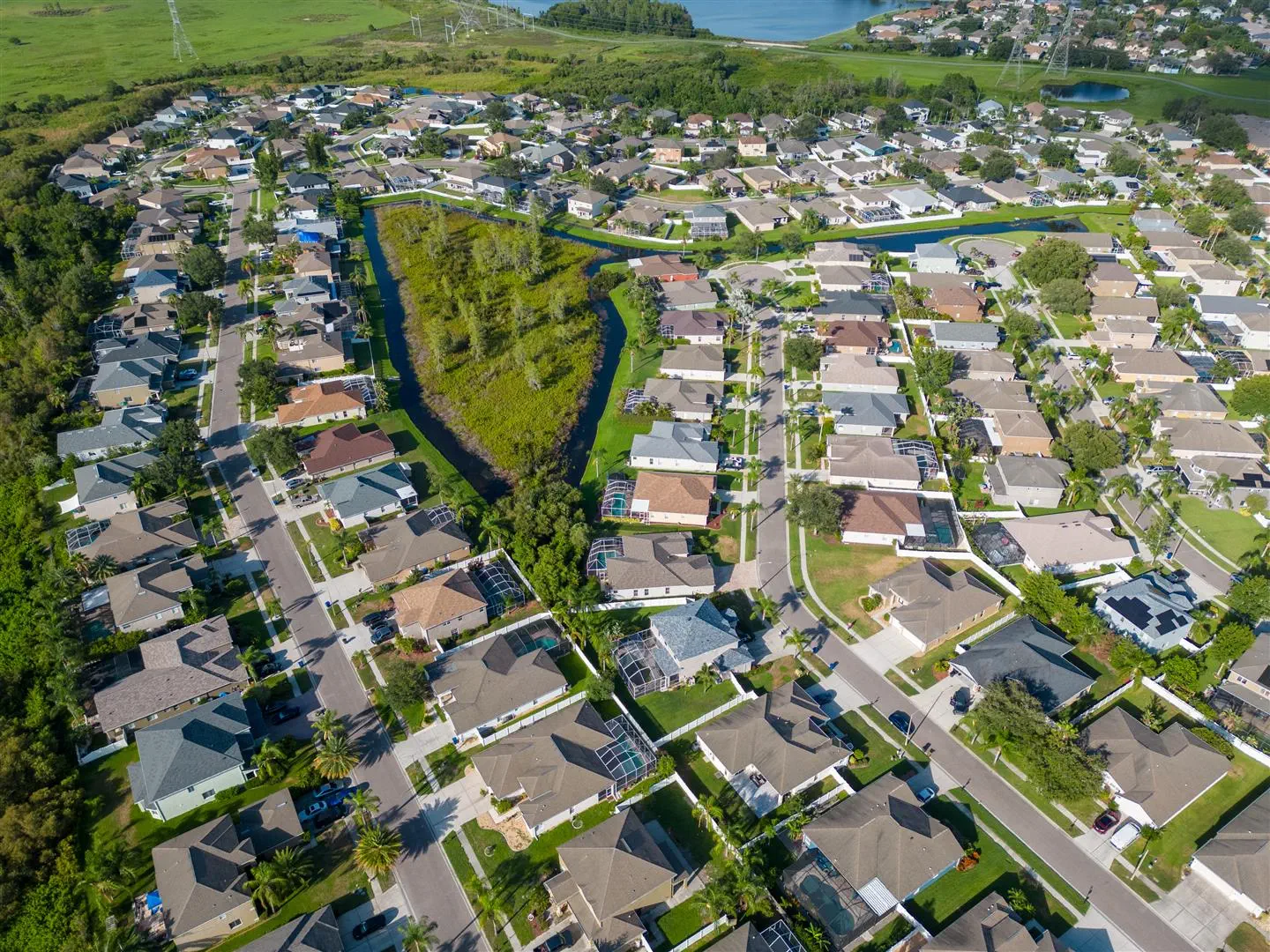 Aerial view of a suburban neighborhood with neat rows of houses, green lawns, and a central canal surrounded by trees, conveying a serene atmosphere.