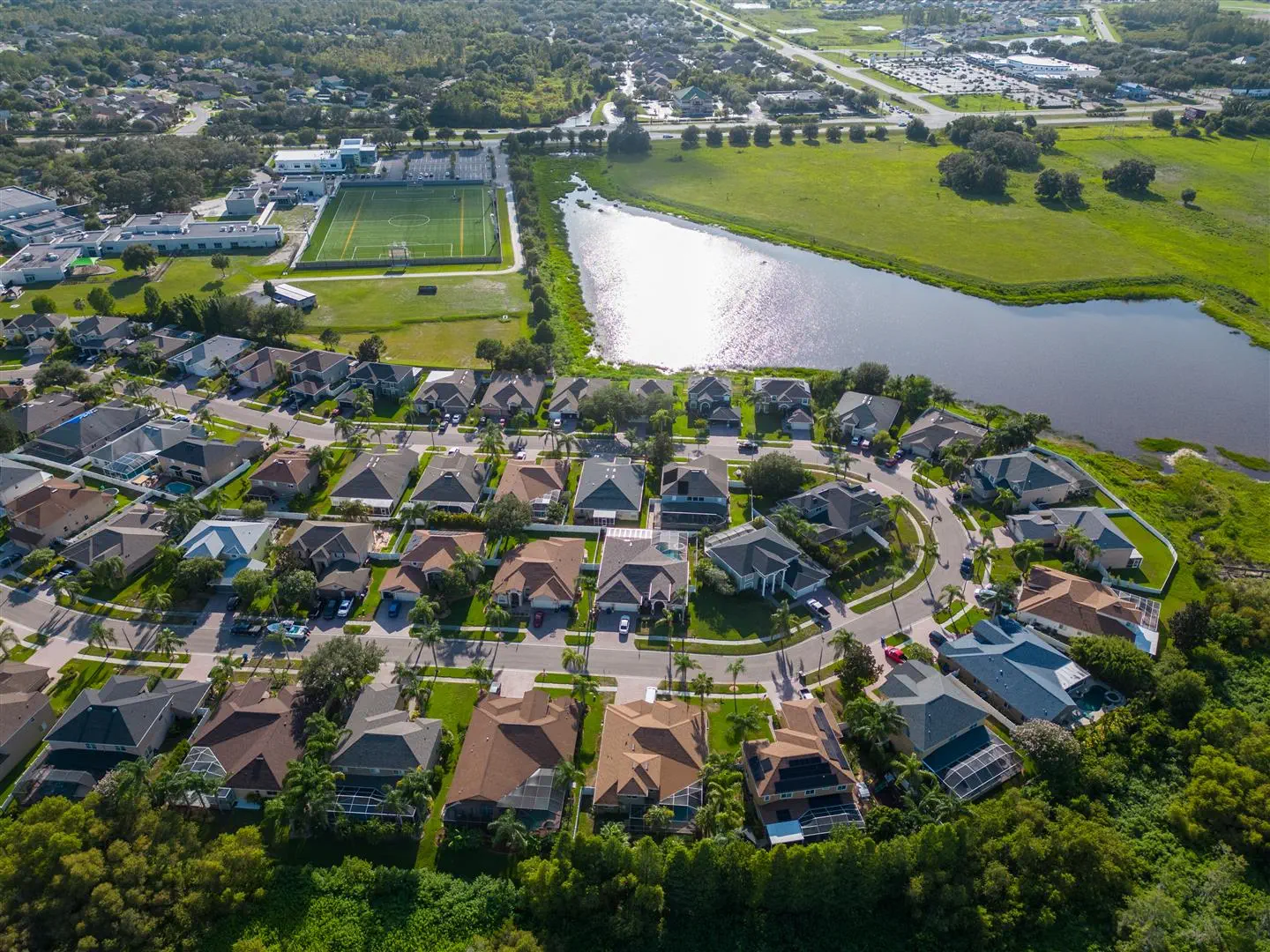 Aerial view of a suburban neighborhood with neatly arranged houses and cul-de-sacs. A lush green field, sports complex, and serene pond are visible.