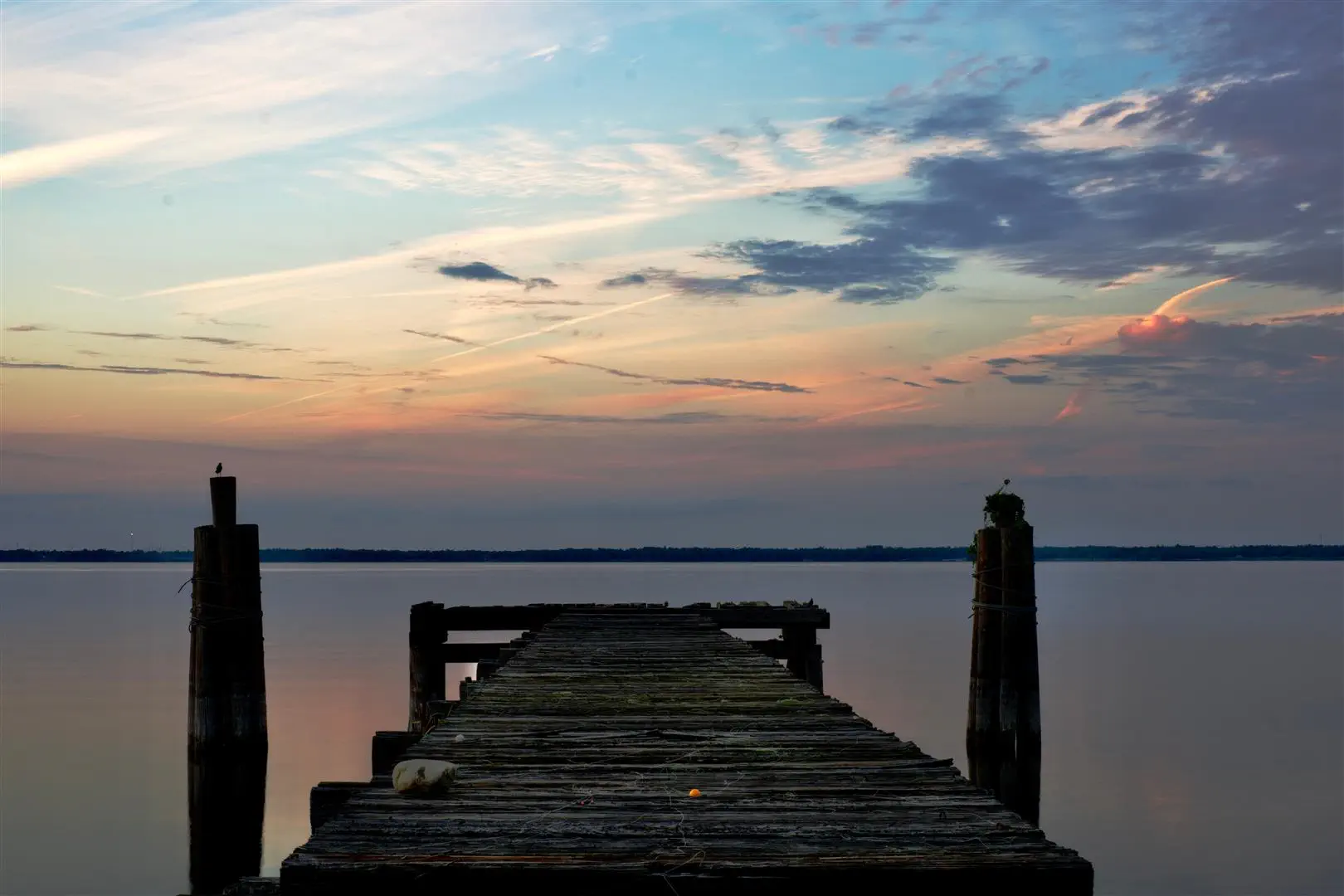 Old wooden pier extending over calm water, under a colorful twilight sky with pastel pink and blue hues. Peaceful and serene atmosphere.