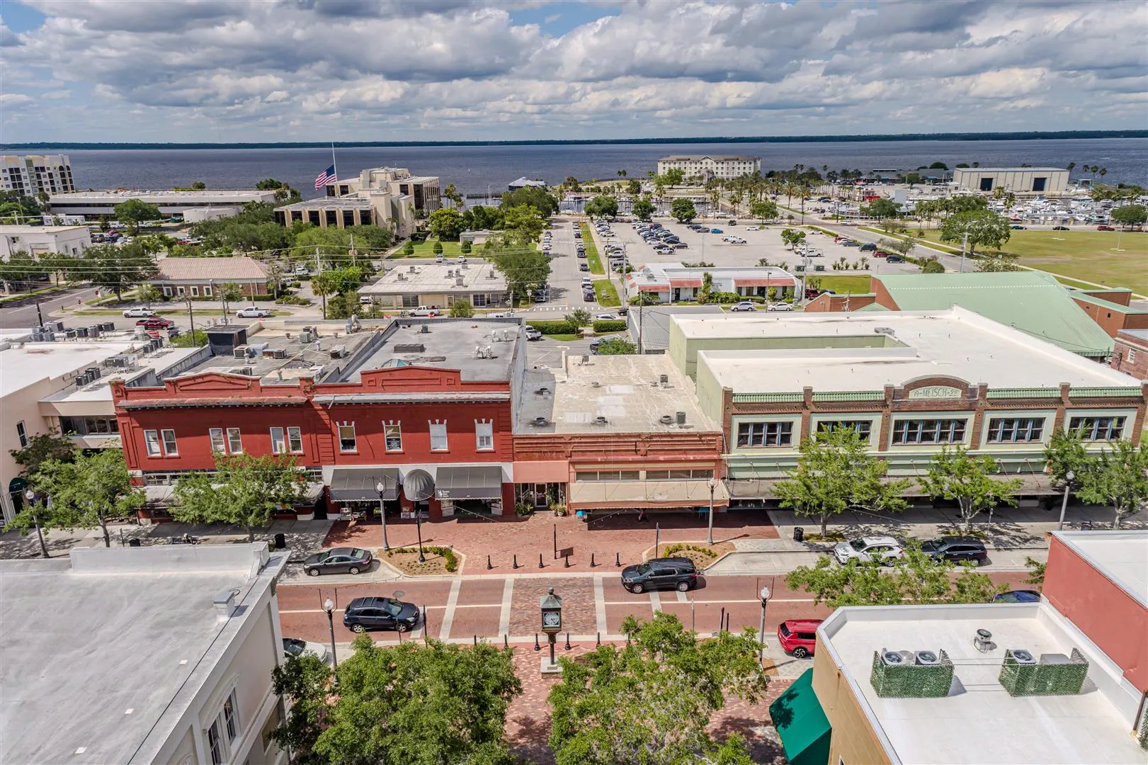 Aerial view of a small town with colorful historic buildings lining a brick street. Cars are parked along the road, and a serene body of water is visible in the background under a partly cloudy sky.