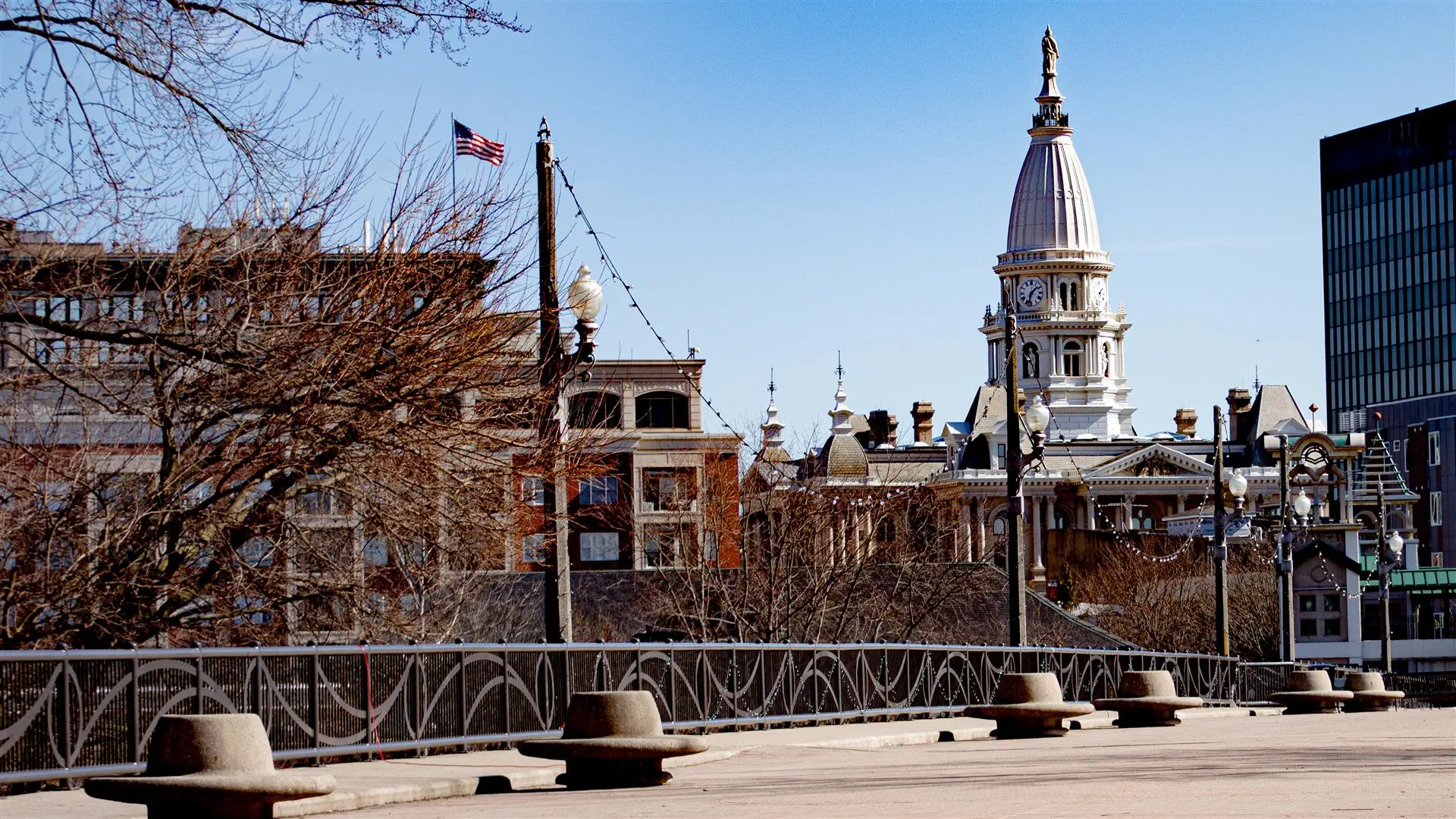 A scenic view of a historic urban skyline featuring an ornate courthouse with a tall dome. Bare trees and a flag add character under a clear blue sky.
