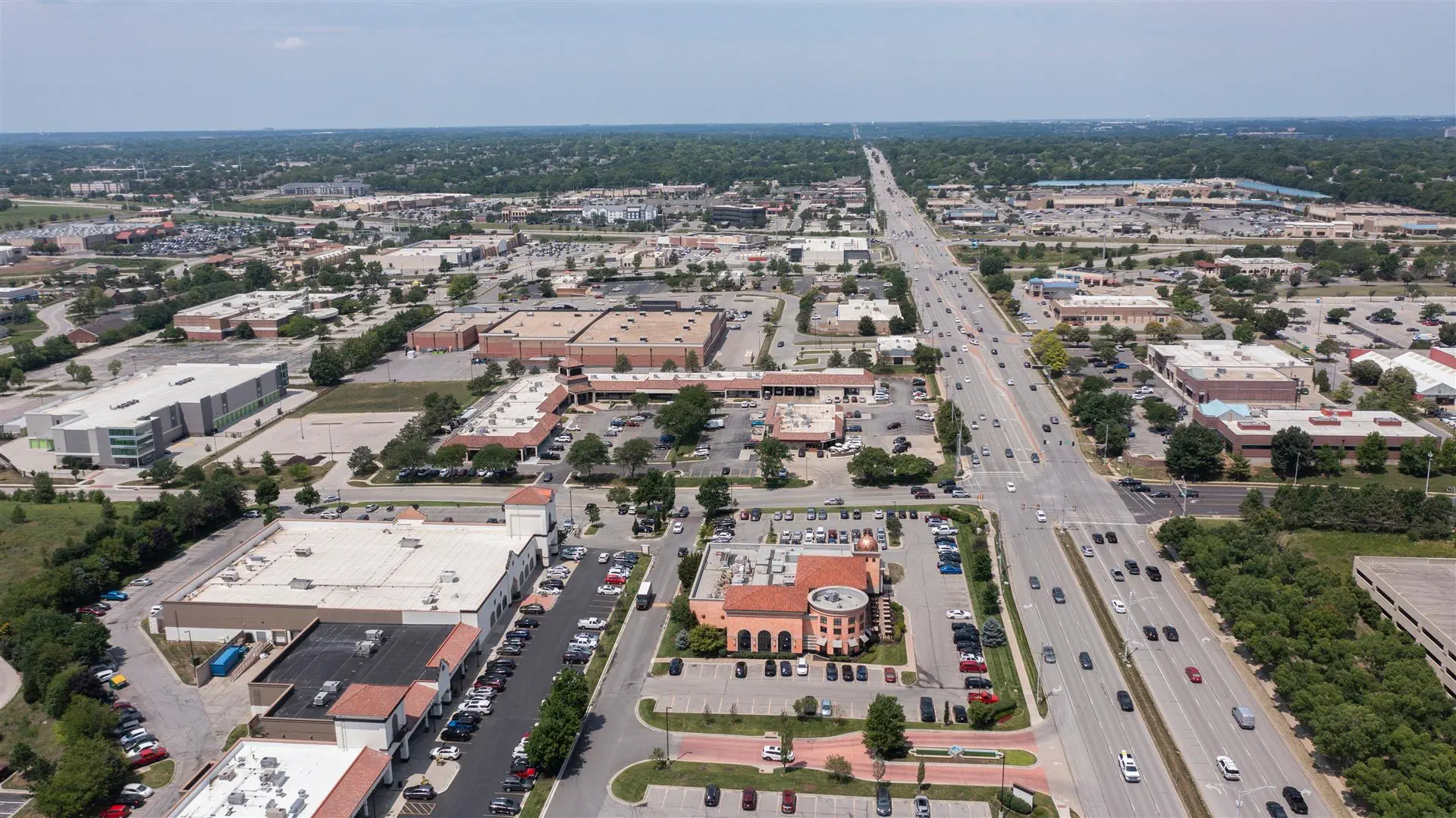 Aerial view of a suburban area with a major road lined by commercial buildings and parking lots. The scene conveys a busy, developed neighborhood.