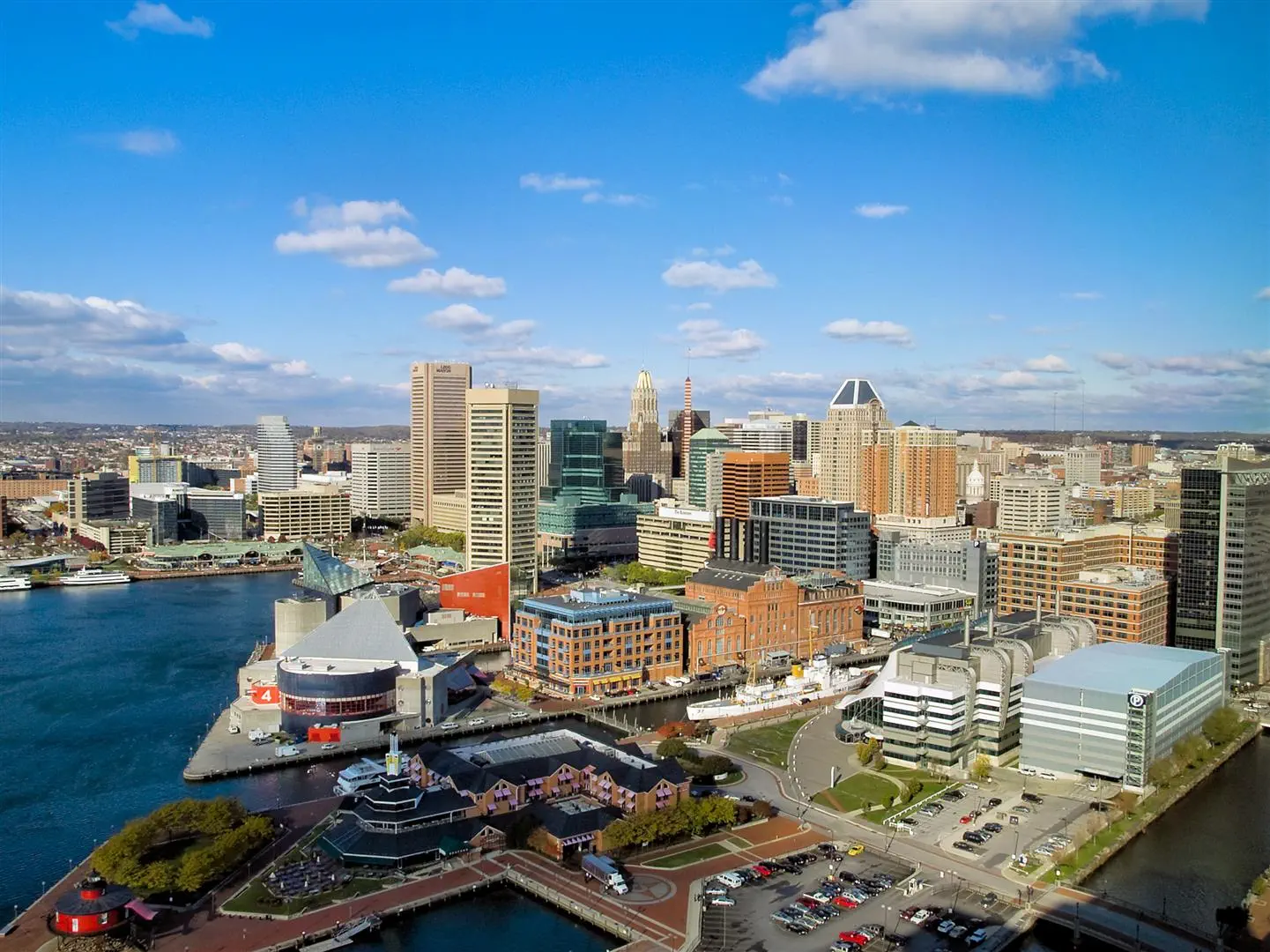 A vibrant cityscape of Baltimore featuring modern skyscrapers, historic brick buildings, and a waterfront. Bright blue sky with scattered clouds creates an inviting atmosphere.