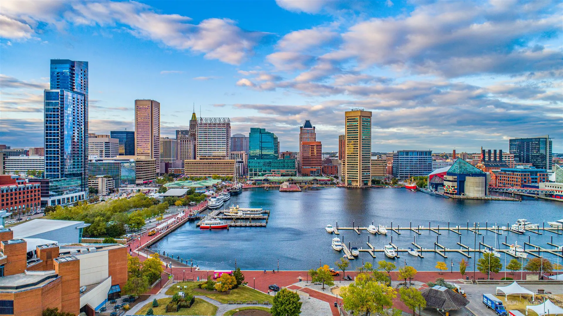 A vibrant cityscape of Baltimore's Inner Harbor under a partly cloudy sky. Skyscrapers border the harbor, reflecting in the water dotted with boats.
