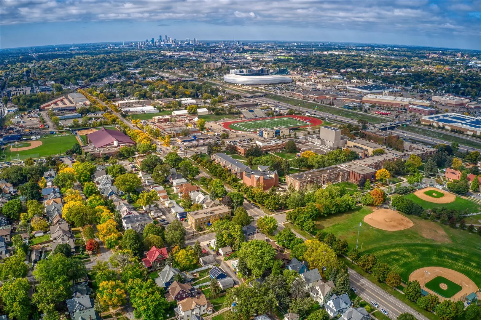 Aerial view of a suburban neighborhood with tree-lined streets, houses, sports fields, and a large stadium. The city skyline is visible on the horizon.
