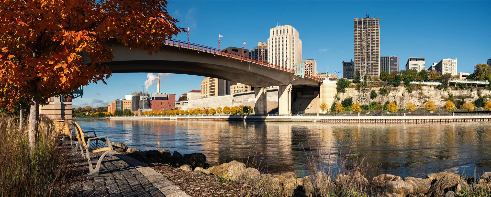A calm river scene with a bridge spanning across it. On the left, autumnal trees and a park bench are by the shoreline. In the background, a city skyline with tall buildings under a clear blue sky.