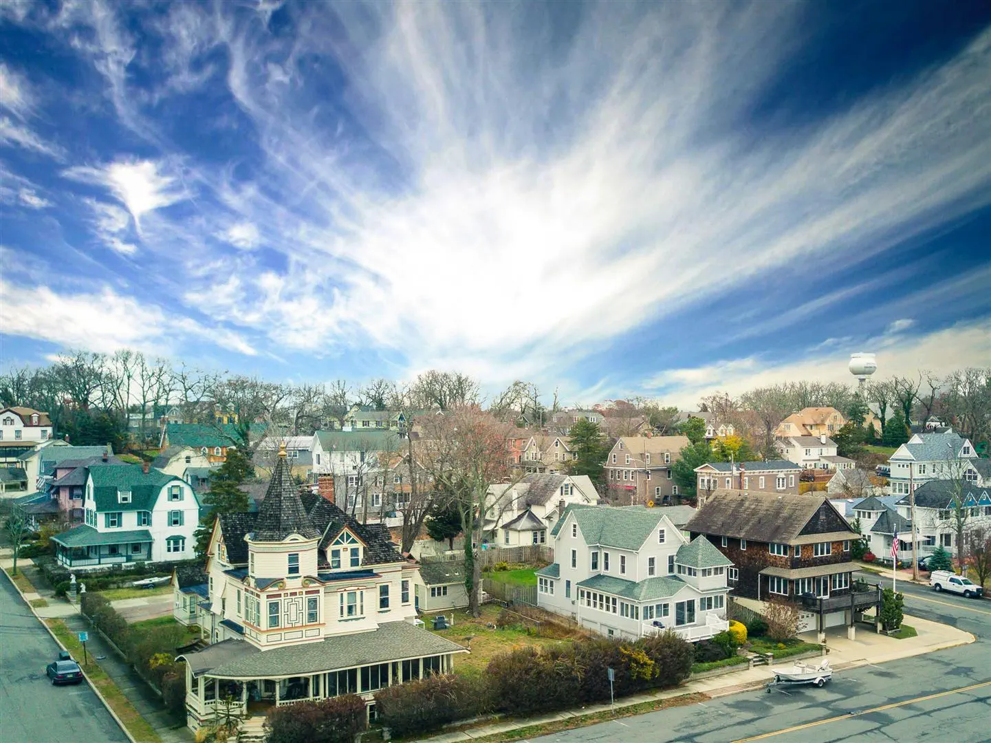 Charming neighborhood with Victorian and modern houses under a vivid blue sky with wispy clouds. A peaceful, suburban atmosphere is conveyed.