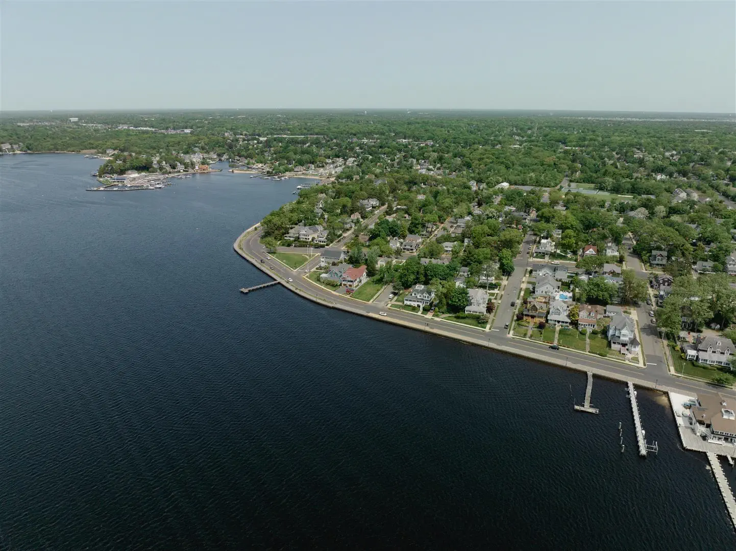 Aerial view of a coastal town with a curved shoreline and docks. Lush green trees surround houses, creating a peaceful, serene atmosphere.