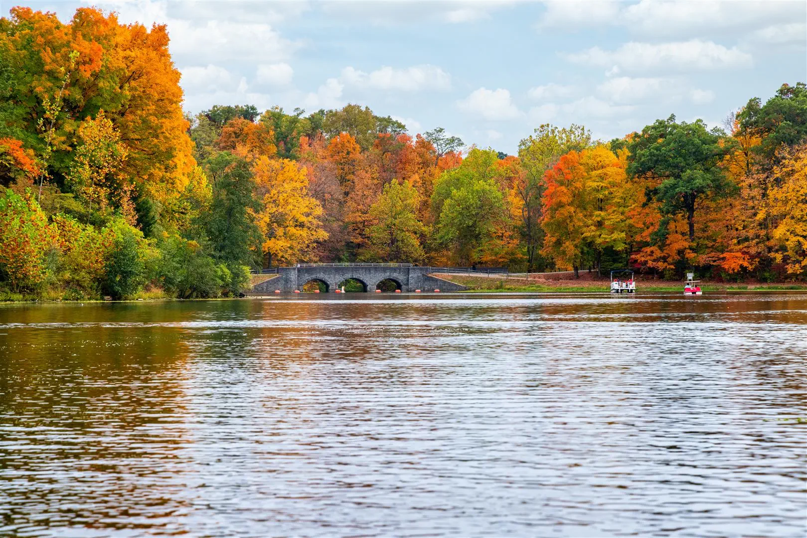 A serene lake reflects vibrant autumn foliage in shades of orange, yellow, and green. A stone bridge spans the water, with small boats floating nearby.