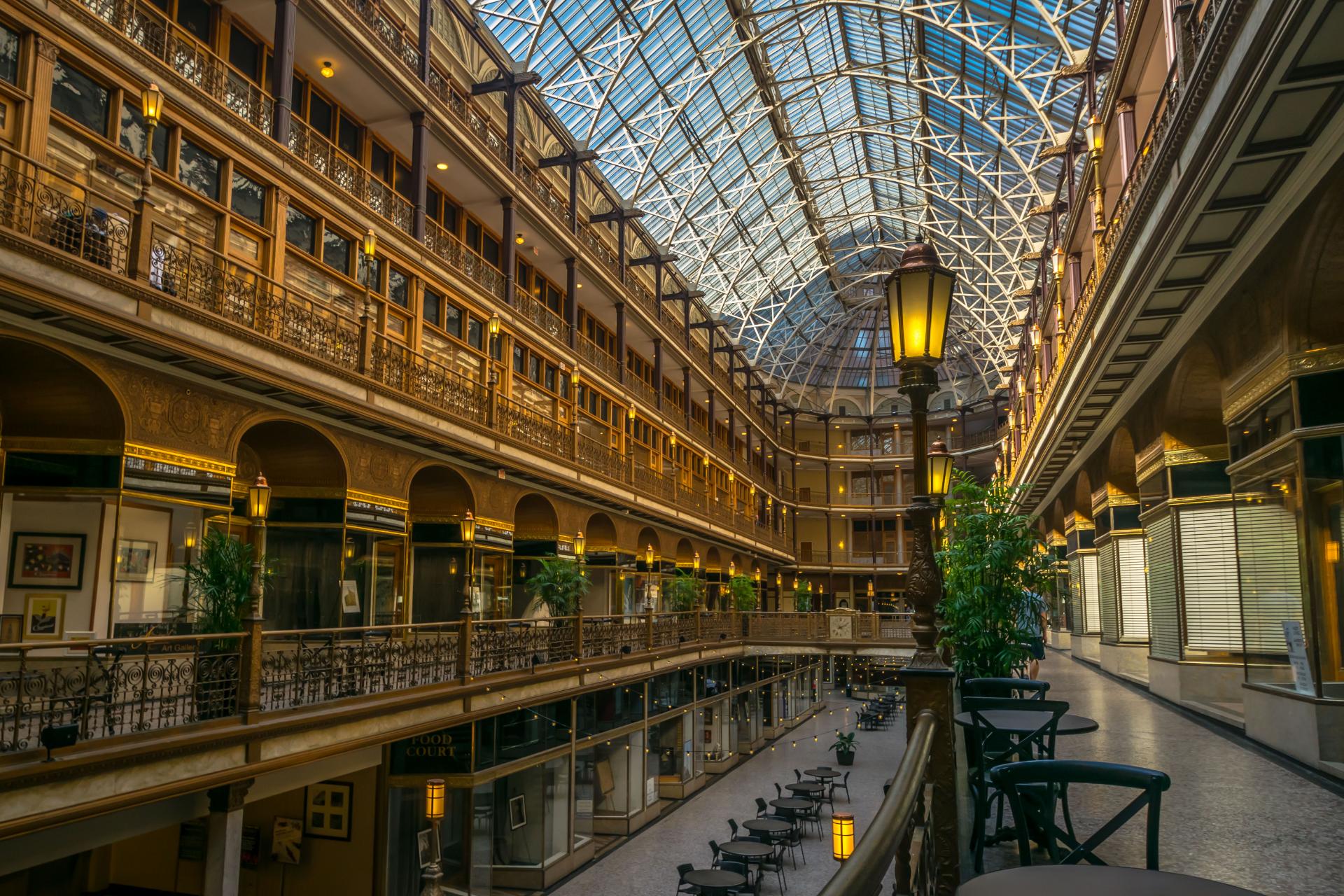 Elegant indoor shopping arcade with a high glass dome ceiling, ornate metal railings, warm lighting, and potted plants. Tables line the floor's edge.