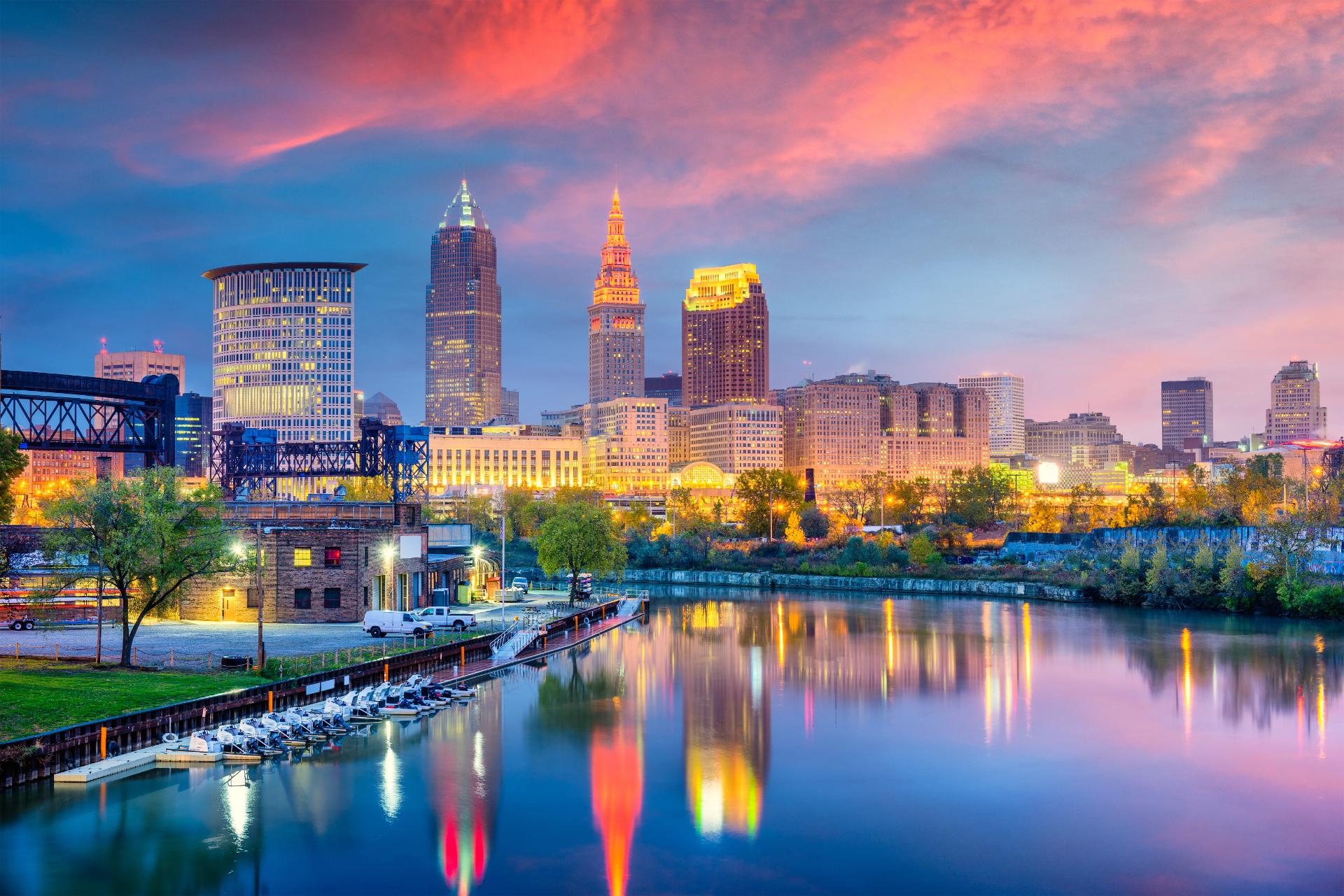 Colorful urban skyline of a city at sunset, reflected in a calm river. Buildings are illuminated, and the sky is a vibrant pink and blue, creating a serene atmosphere.
