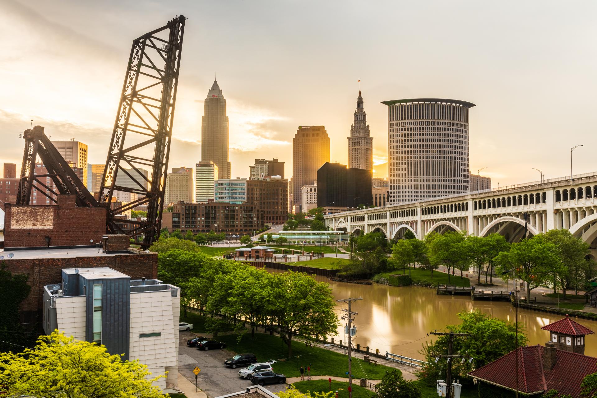 Skyline of a city at sunset with tall buildings and a bridge over a river. A lifted crane in the foreground and lush green trees add contrast.
