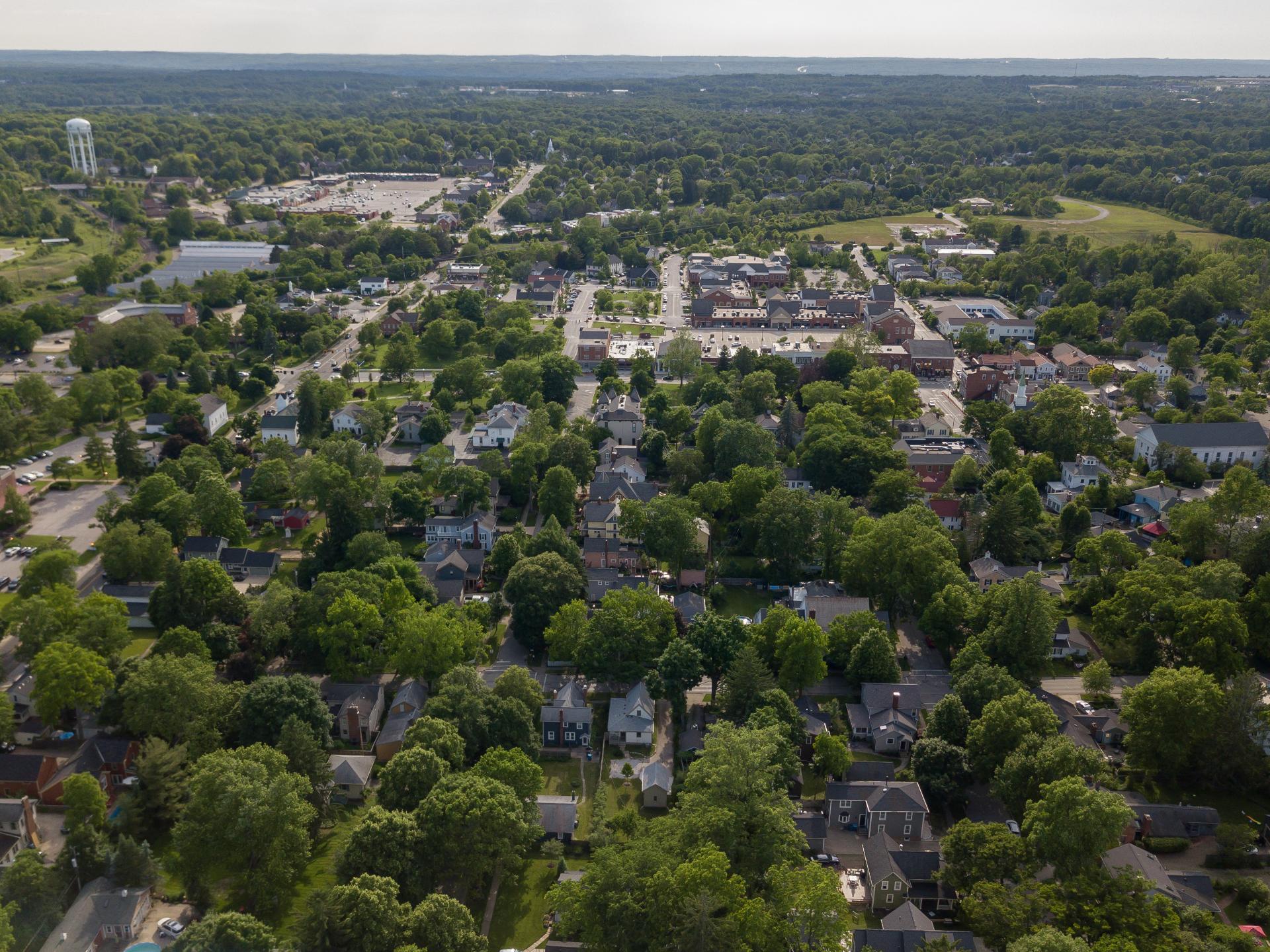 Aerial view of a suburban neighborhood with tree-lined streets, houses, and small buildings. A water tower and commercial area are visible in the distance.
