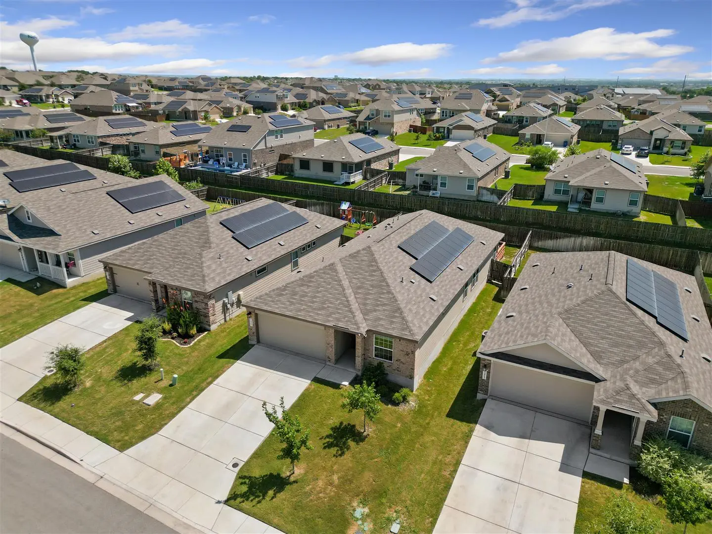 Suburban neighborhood with rows of single-story homes, each with solar panels on the roofs. Neatly maintained lawns and clear blue sky overhead.