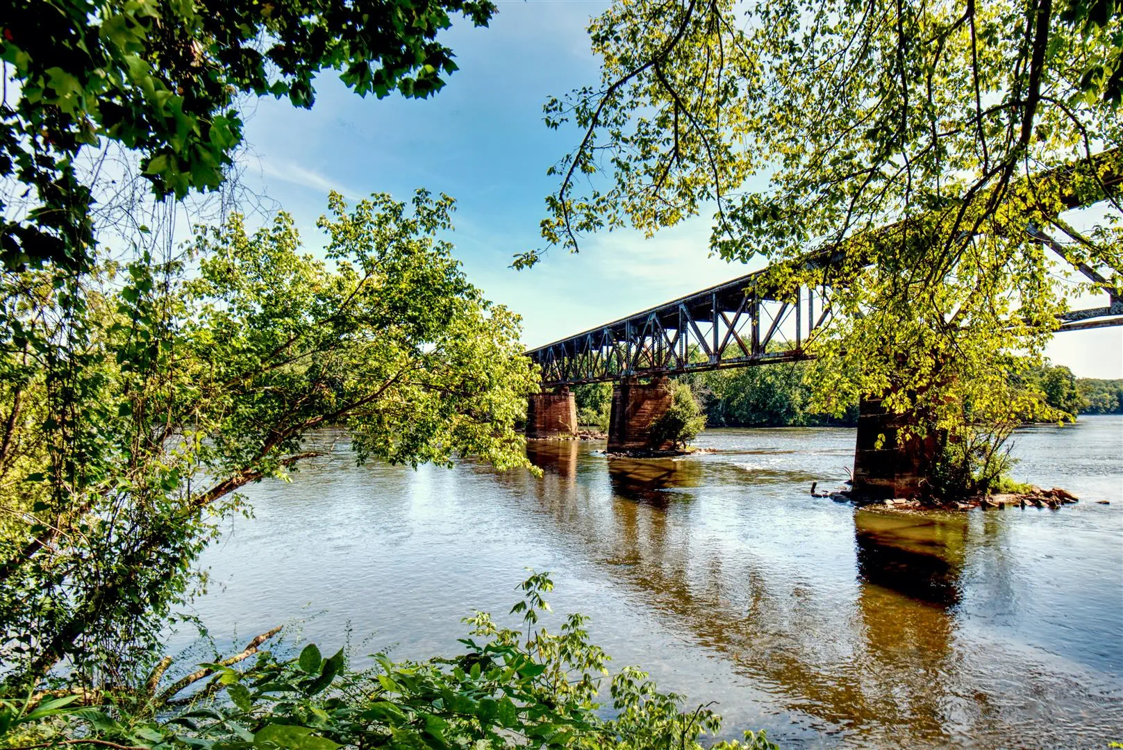 Railway bridge spans a tranquil river, surrounded by lush green trees. Sunlight filters through the leaves, creating reflections on the water. Peaceful scene.
