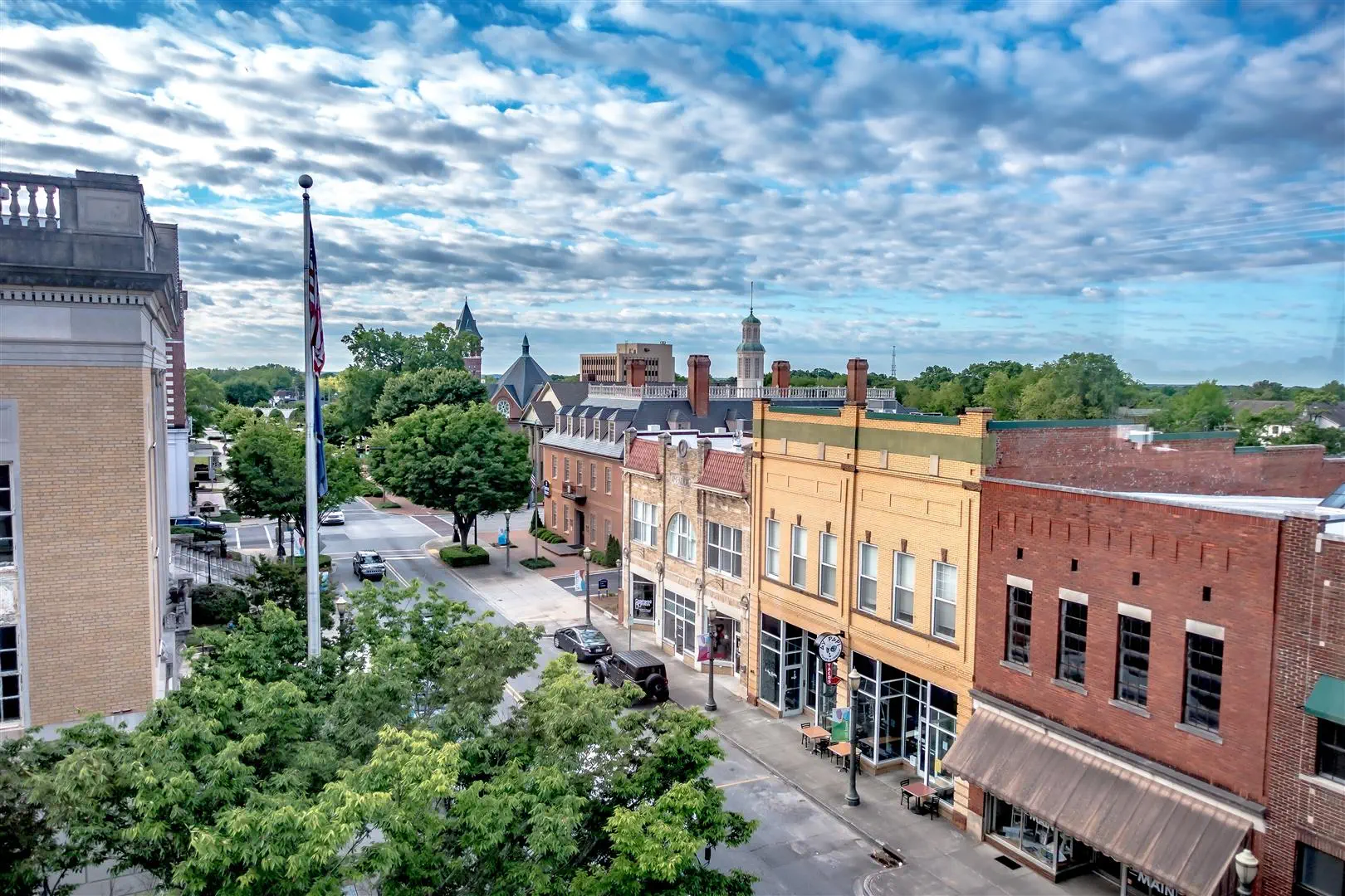 Aerial view of a quaint downtown street with historic buildings. A tree-lined square and an American flag create a serene atmosphere under a cloud-dotted sky.