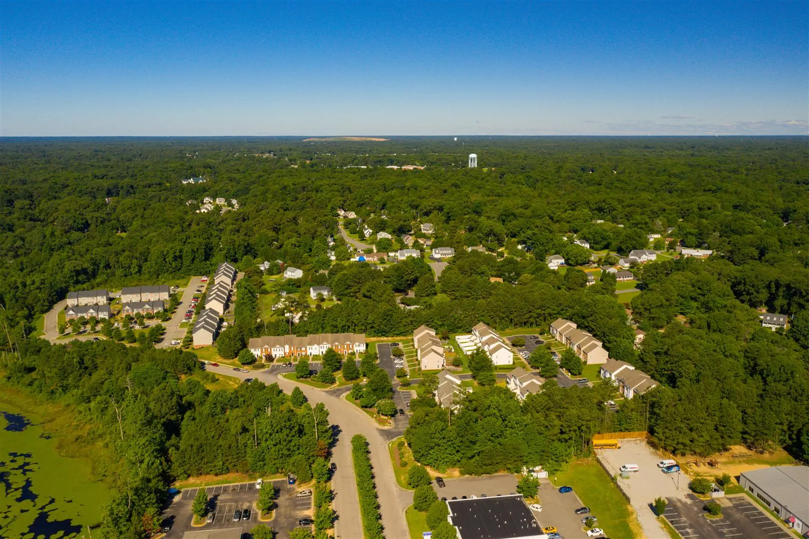Aerial view of a suburban neighborhood surrounded by dense green forests under a clear blue sky. Rows of houses and roads are visible, creating a peaceful, organized landscape.