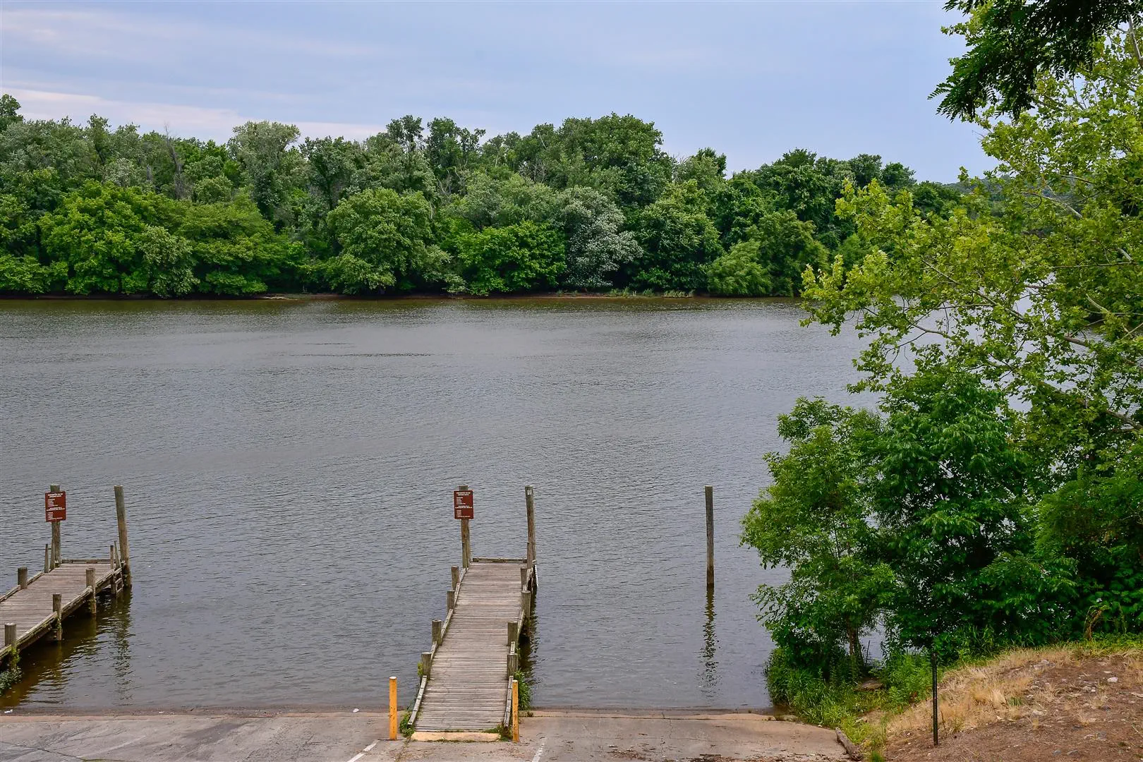 Wooden docks extend into a calm river surrounded by lush green trees under a cloudy sky, creating a serene and peaceful atmosphere.