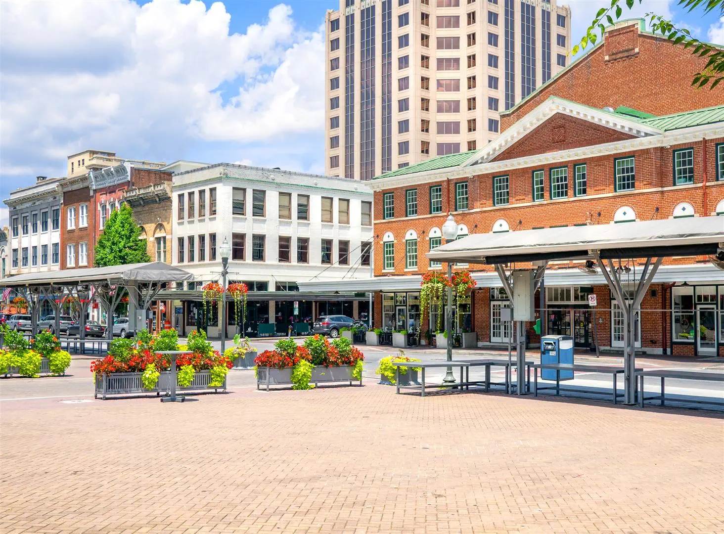 A lively urban scene with historic brick and white buildings under a blue sky. Colorful planters line the plaza, conveying a vibrant, welcoming atmosphere.
